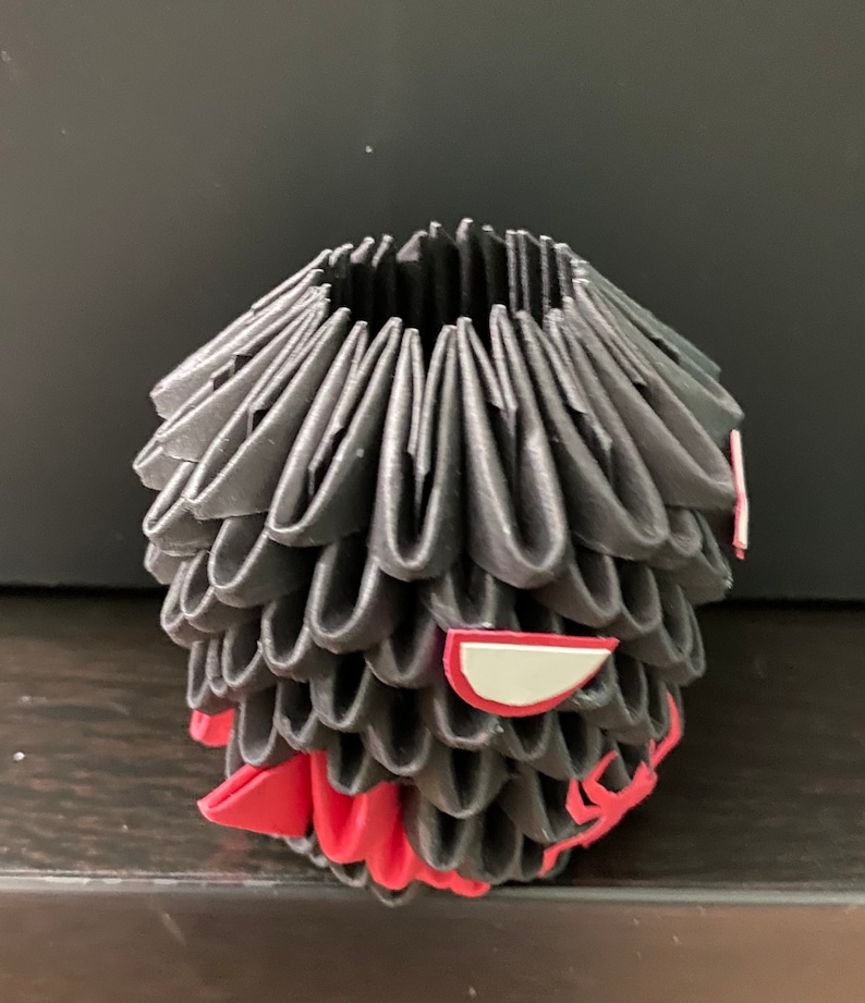 Może przedstawiać: Czarna rzeźba origami z czerwonymi i białymi akcentami. Rzeźba ma kształt głowy ze stylizowaną twarzą.