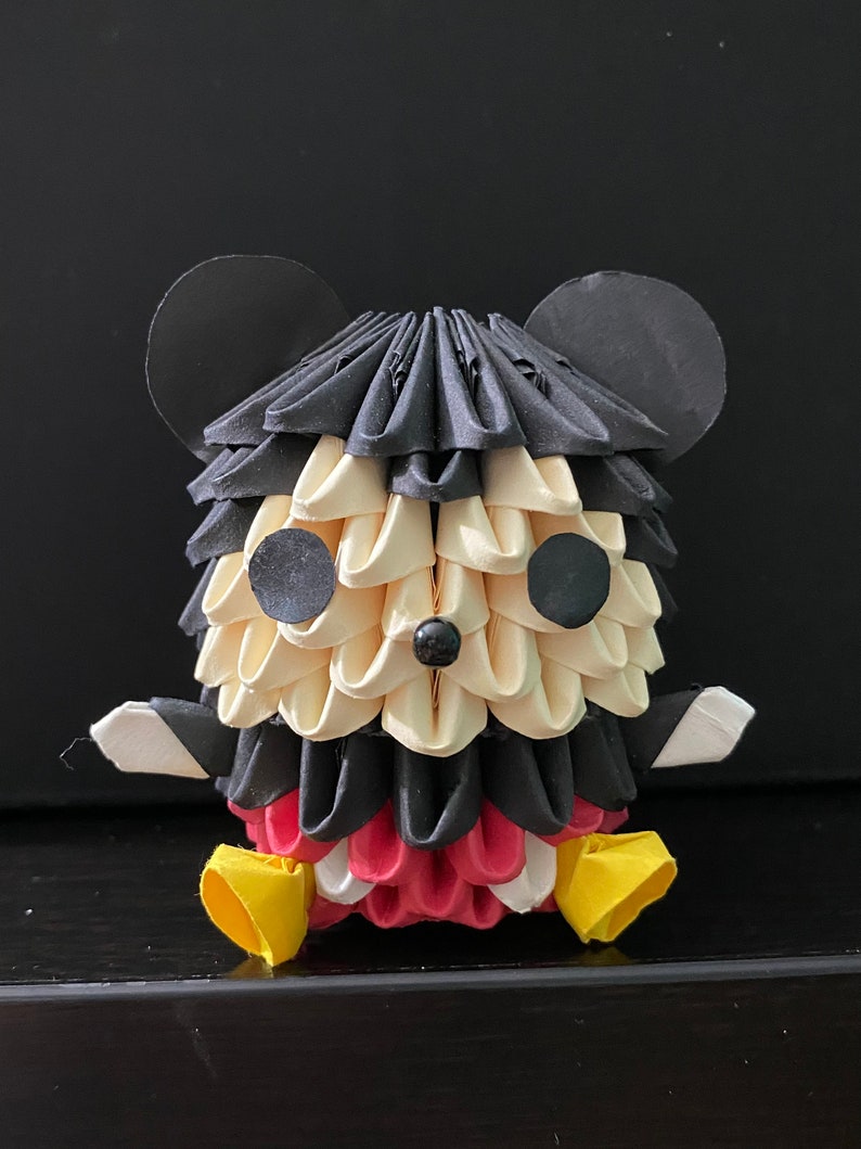 May include: An origami Mickey Mouse figure made from black, white and red paper. The figure is sitting with its legs crossed and has large black ears.