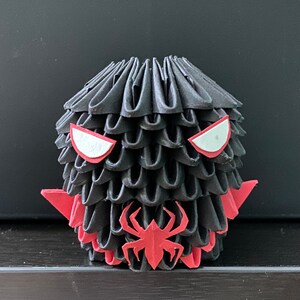 Może przedstawiać: Czarna figurka origami z czerwonymi oczami i czerwonym wzorem pająka. Figurka jest wykonana ze złożonego papieru i ma fakturowaną powierzchnię.