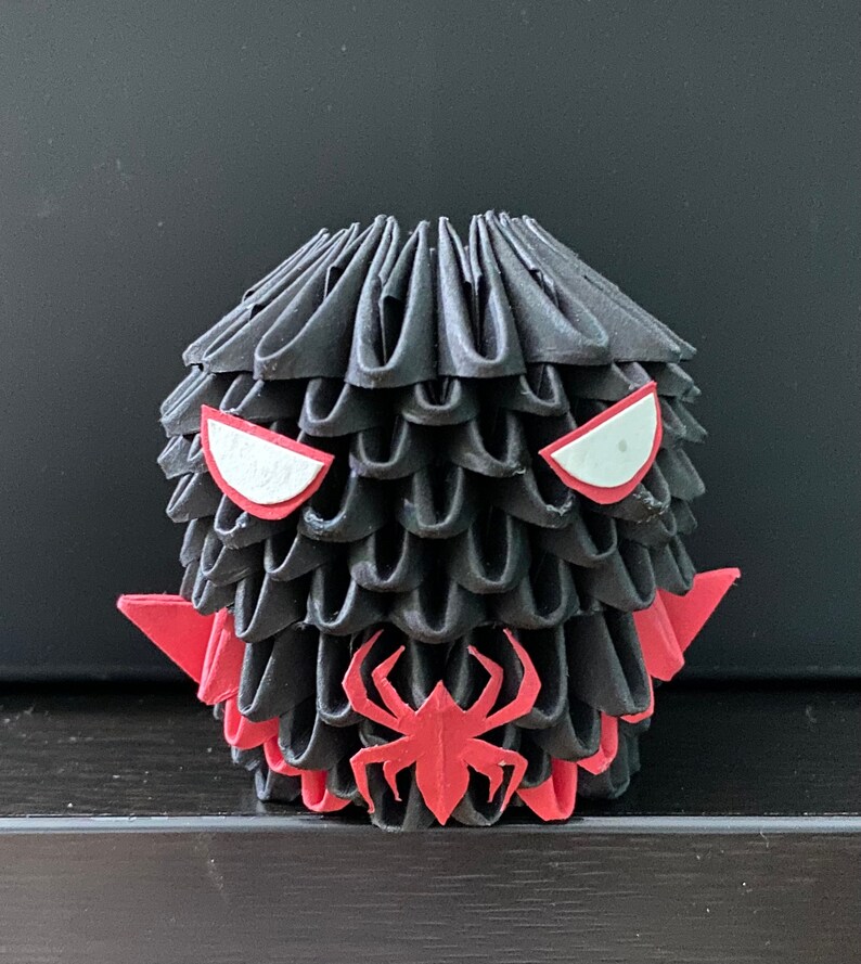 Może przedstawiać: Czarna figurka origami z czerwonymi oczami i czerwonym wzorem pająka. Figurka jest wykonana ze złożonego papieru i ma fakturowaną powierzchnię.