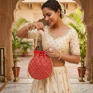 May include: A vibrant red drawstring pouch bag featuring an intricate gold-toned grid pattern with small white bead accents. The bag has a pearl-beaded handle and gold-toned drawstring cords with decorative tassels at the ends, held by a person.