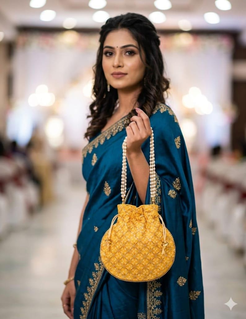 May include: A round yellow drawstring pouch bag featuring a repeating white floral embroidery pattern and a beaded handle made of multiple strands of small white pearls. The bag has a cinched top closure with matching yellow fabric drawstrings.