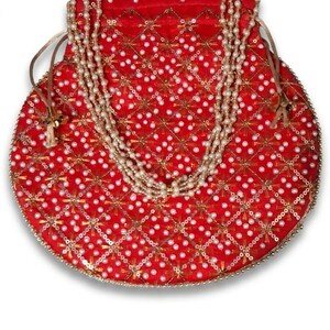 May include: A round red drawstring pouch featuring a gold-toned lattice pattern with embedded white faux pearls. The bag is adorned with a decorative handle made of multiple strands of white faux pearls and includes thin side drawstrings.
