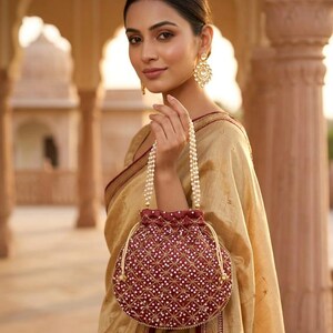 May include: A deep maroon drawstring pouch bag featuring a geometric pattern of white beads and gold thread embroidery. The bag has a handle made of two strands of white pearls and a drawstring closure with gold-toned cord ends and matching beads.