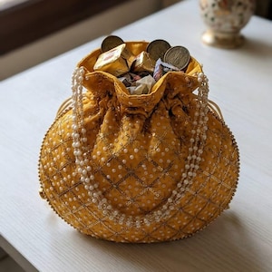 May include: A mustard yellow drawstring pouch bag featuring a quilted diamond pattern with gold metallic thread, small white faux pearls, and a beaded handle. The bag is filled with several gold-wrapped square candies and round metal coins.