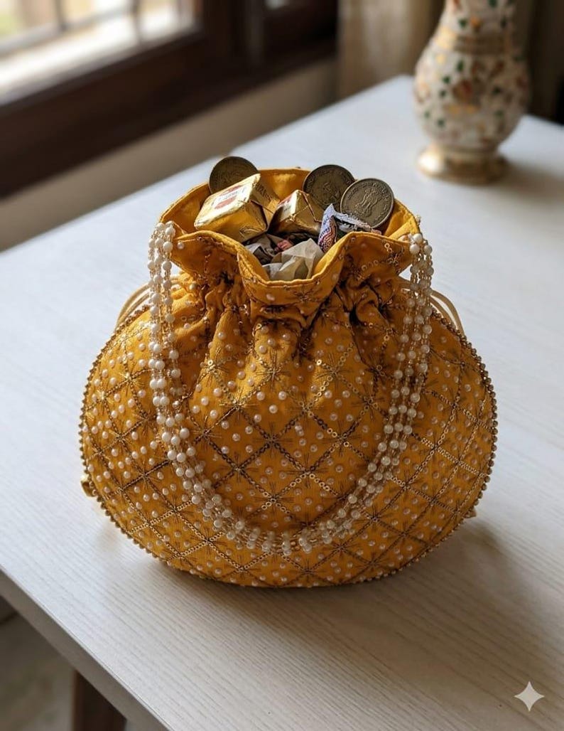 May include: A mustard yellow drawstring pouch bag featuring a quilted diamond pattern with gold metallic thread, small white faux pearls, and a beaded handle. The bag is filled with several gold-wrapped square candies and round metal coins.