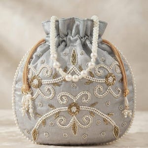 Könnte beinhalten: Ein silberner Beutel mit Kordelzug, verziert mit aufwendigen Stickereien aus weißen Perlen und goldenem Metallic-Garn. Die Tasche hat einen gerafften Verschluss, einen Perlenhenkel und goldene Kordeln mit perlenbesetzten Quasten an den Seiten.