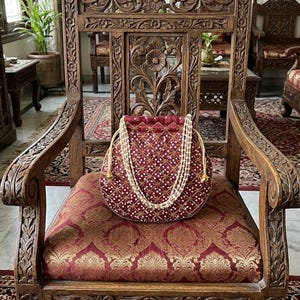 May include: A deep red drawstring pouch featuring a repeating gold embroidered star pattern with small white beads at each intersection. The bag includes a double-strand white pearl handle and gold-toned drawstrings, creating a structured circular shape.