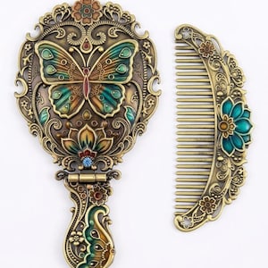 Puede incluir: Un espejo de mano y un peine a juego con acabado en bronce antiguo. El espejo presenta un motivo de mariposa y flores con detalles en esmalte verde azulado, verde y naranja, mientras que el peine muestra un diseño floral coordinado.