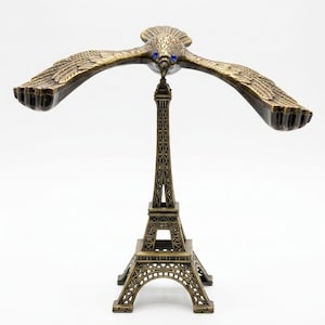 May include: A bronze-colored metal sculpture featuring a bird with outstretched wings balanced on the tip of a miniature Eiffel Tower replica. The bird has small, bright blue circular accents for eyes, and the entire structure displays intricate lattice detailing on the tower base.
