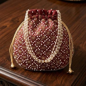 May include: A deep red drawstring pouch bag featuring an intricate pattern of gold metallic thread embroidery and small white faux pearls. The bag includes a long, multi-strand white pearl handle and light beige drawstring cords with gold tips.