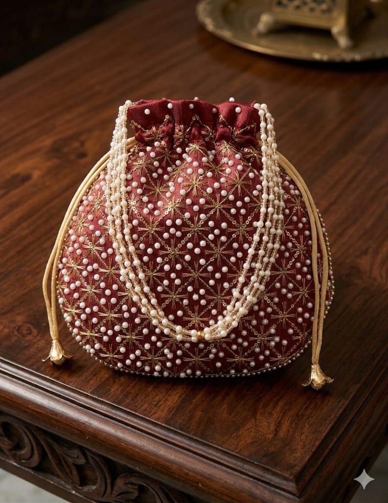 May include: A deep red drawstring pouch bag featuring an intricate pattern of gold metallic thread embroidery and small white faux pearls. The bag includes a long, multi-strand white pearl handle and light beige drawstring cords with gold tips.