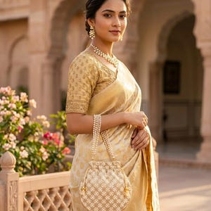 May include: A round, cream-colored drawstring handbag featuring a detailed gold-toned lattice embroidery pattern and a handle made of multiple strands of small white pearls, designed with a gathered top closure and matching gold-toned tassels.