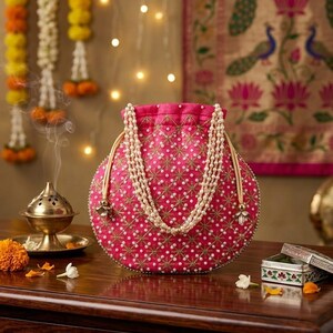 May include: A bright pink drawstring pouch bag featuring intricate gold embroidery and small white pearl embellishments. The bag has a double-strand pearl handle and a gathered top closure, showcasing a detailed star-like pattern across the fabric.