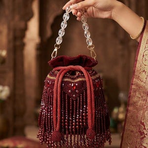 May include: A deep maroon velvet drawstring handbag featuring intricate beadwork, red crystal accents, and a long fringe of matching maroon beads. The bag includes a clear, faceted acrylic bead handle attached with gold-toned metal clasps.