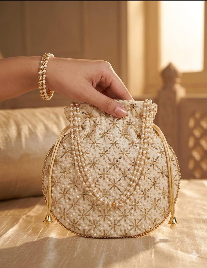 May include: A round, cream-colored drawstring pouch featuring intricate gold embroidery and small white pearl embellishments. The bag includes a handle made of multiple strands of white pearls and gold-toned metal drawstring ends.