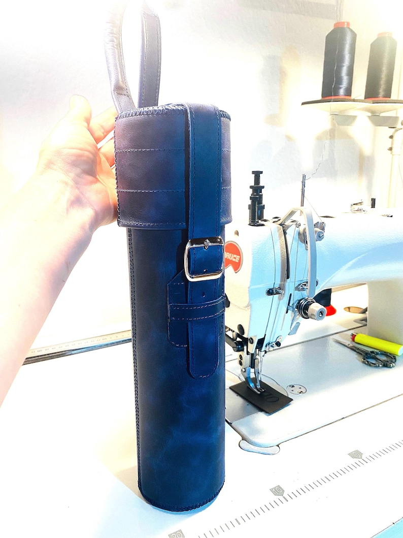 May include: A dark blue leather carrying case with a strap and buckle. The case is cylindrical and has a stitched design. The case is being held by a hand in front of a sewing machine.