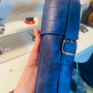 May include: A dark blue leather case with a strap and buckle closure. The case has a stitched design and is made of a textured leather.