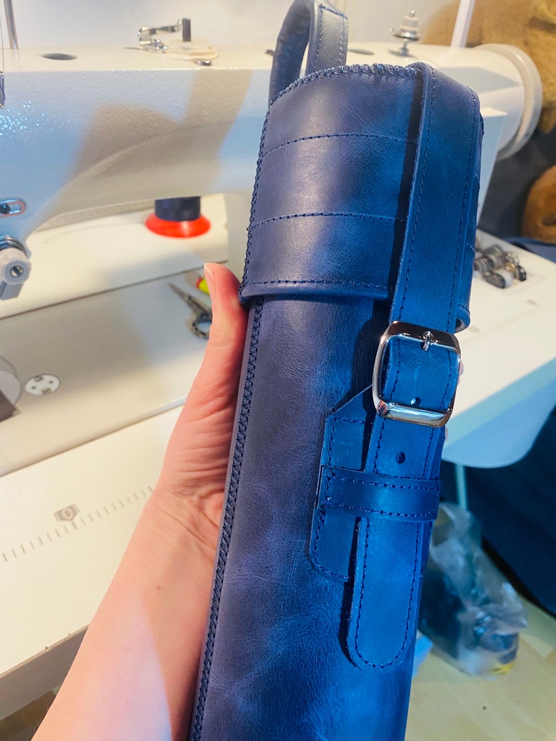 May include: A dark blue leather case with a strap and buckle closure. The case has a stitched design and is made of a textured leather.
