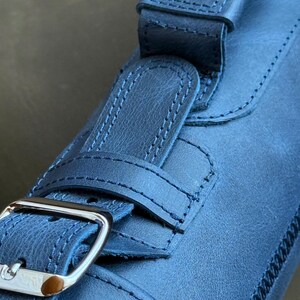 May include: A close-up of a dark blue leather strap with a silver buckle. The strap has a stitched design and is folded over itself.