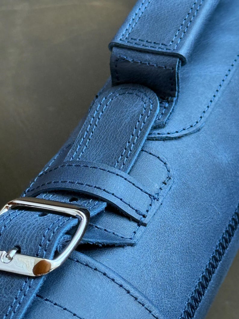 May include: A close-up of a dark blue leather strap with a silver buckle. The strap has a stitched design and is folded over itself.
