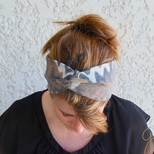 Op de afbeelding: Een bruin, grijs en wit gestreept hoofdband met een gedraaide knoop in het midden. Het hoofdband wordt gedragen door een persoon met bruin haar.