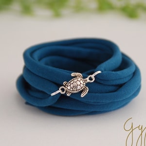 Könnte beinhalten: Ein blaues Stoffarmband mit einem silbernen Schildkrötenanhänger. Das Armband besteht aus mehreren Stofflagen und wird mit einem silbernen Verschluss gebunden.