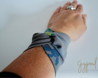 Può includere: Un braccialetto in tessuto grigio con un motivo floreale blu e verde. Il braccialetto è annodato al polso. Il testo "Gypsoul accessories" è visibile nell'angolo in basso a destra dell'immagine.