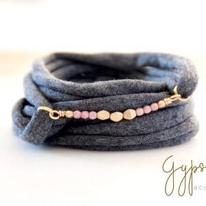 Puede incluir: Un brazalete de tela gris con un acento de cuentas doradas y rosadas. El brazalete está envuelto alrededor de sí mismo varias veces y tiene un cierre dorado.