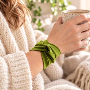 Peut inclure: Un bracelet en tissu vert olive vif présentant un nœud torsadé central. L'accessoire est fabriqué dans une matière souple et flexible qui s'enroule confortablement autour du poignet, créant un aspect superposé avec une texture lisse.