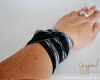 Può includere: Un braccialetto in tessuto a righe bianche e nere. Il braccialetto è annodato e indossato al polso. Il testo "Gypsoul accessories" è visibile nell'angolo in basso a destra dell'immagine.