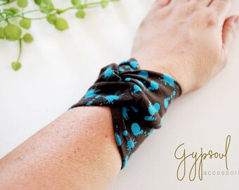 Può includere: Un braccialetto in tessuto marrone con un motivo a schizzi di vernice blu. Il braccialetto è annodato e indossato al polso. Il testo "Gypsoul accessories" è visibile nell'angolo in basso a destra dell'immagine.