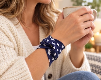 Può includere: Un braccialetto in tessuto blu scuro caratterizzato da un motivo ripetuto di piccole stelle bianche. Il braccialetto è progettato con un dettaglio a nodo intrecciato al centro ed è indossato al polso, mostrando una struttura tessile morbida.