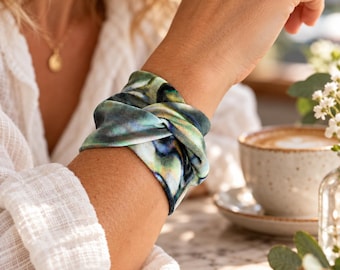 Può includere: Un ampio bracciale in tessuto caratterizzato da un motivo iridescente vorticoso nei toni del blu profondo, verde smeraldo e oro metallizzato. Il materiale è arricciato e attorcigliato per creare un aspetto stratificato e strutturato.