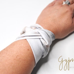 Può includere: Un braccialetto in tessuto bianco e grigio con un design annodato. Il braccialetto è indossato al polso di una persona. Il testo "Gypsoul accessories" è visibile nell'angolo in basso a destra dell'immagine.