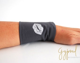 Può includere: Un braccialetto da polso grigio con un design a labbra con glitter argentato. Il braccialetto è realizzato in un tessuto morbido ed elastico.
