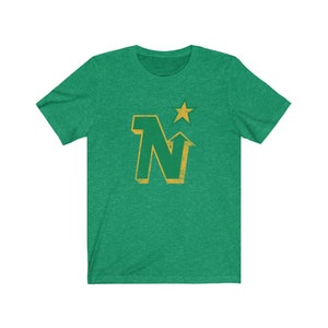 Peut inclure: T-shirt vert avec un motif dor&eacute; vieilli. Le motif repr&eacute;sente la lettre "N" avec une fl&egrave;che vers le haut et une &eacute;toile au-dessus. Le t-shirt est fait d'une mati&egrave;re douce.