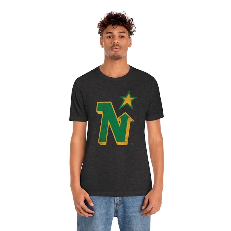 Peut inclure: T-shirt gris fonc&eacute; avec un motif vieilli vert et or en forme de lettre "N" et une &eacute;toile dor&eacute;e. Le t-shirt &agrave; col rond est fait d'une mati&egrave;re douce.