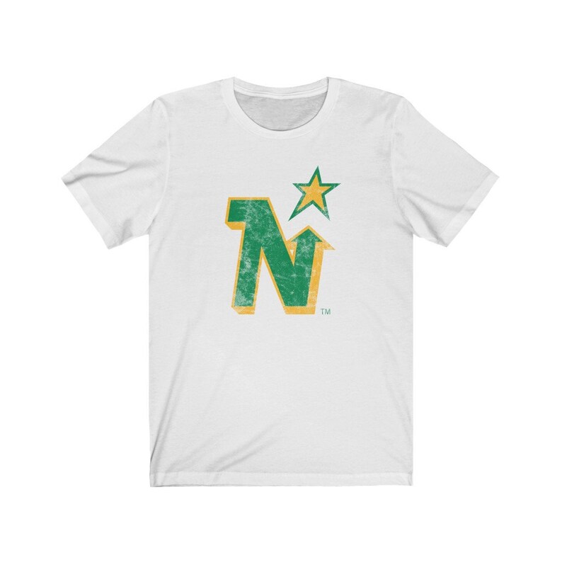 Peut inclure: T-shirt blanc avec un motif vert et or vieilli. Le motif comprend la lettre "N" avec une fl&egrave;che vers le haut et une &eacute;toile. Le t-shirt est fait d'une mati&egrave;re douce.