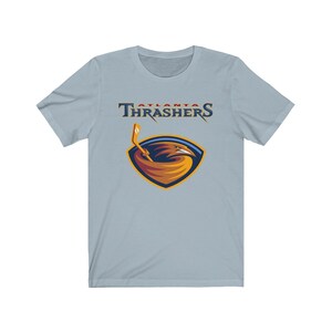 May include: A light blue t-shirt with the Atlanta Thrashers logo. The logo features a red, white, and blue color scheme with a stylized bird holding a hockey stick.