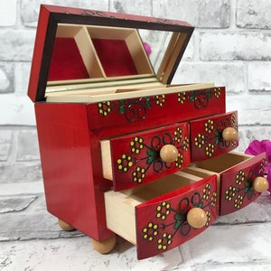 Puede incluir: Caja de joyería de madera roja con un espejo en el interior y cuatro cajones. Los cajones tienen diseños florales pintados y pomos de madera.