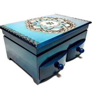 Puede incluir: Una caja de joyería de madera azul con dos cajones y un diseño floral decorativo en la tapa.