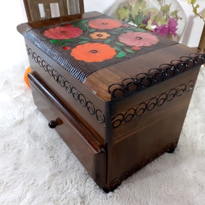 Puede incluir: Una caja de madera con un cajón y un diseño floral en la tapa. Las flores están pintadas en tonos de rojo, naranja y rosa.