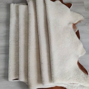 May include: A stack of five white sheepskin rugs with brown leather backing. The rugs are folded in half and stacked on top of each other.