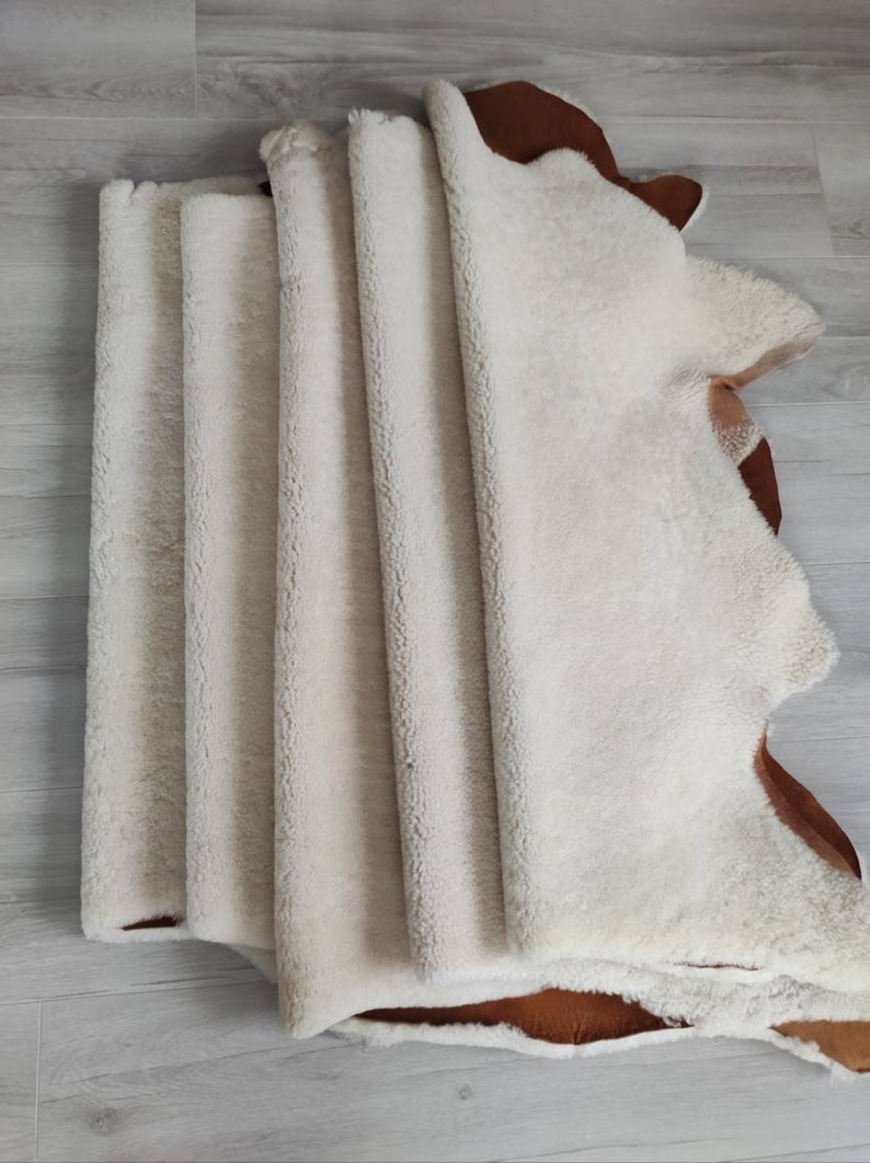 May include: A stack of five white sheepskin rugs with brown leather backing. The rugs are folded in half and stacked on top of each other.