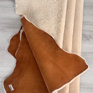 May include: A folded, rectangular rug with a brown leather exterior and a cream-colored, fluffy interior. The rug is made of a soft material and has a natural, organic shape. The rug is displayed on a light gray wooden surface.