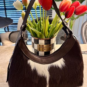 May include: A hobo-style handbag crafted from dark brown and white cowhide with a smooth dark brown leather trim and a single shoulder strap. The bag features a zipper closure along the top edge and a curved silhouette with a textured fur finish.