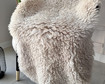 May include: A cream-colored, long-pile sheepskin rug draped over a chair. The texture is thick, curly, and shaggy, covering the seat and backrest of the chair. The chair features black legs with gold-toned metal tips at the base.