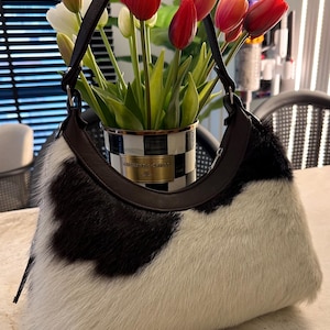 May include: A hobo-style handbag featuring a black and white cowhide pattern with a textured, furry finish. The bag has a dark brown leather handle attached with silver-toned hardware and a curved top opening, showcasing a distinct animal-inspired design.