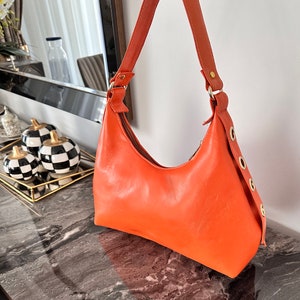 May include: A bright orange leather handbag with a single strap and gold hardware. The bag has a curved shape and features multiple gold grommets on the side.